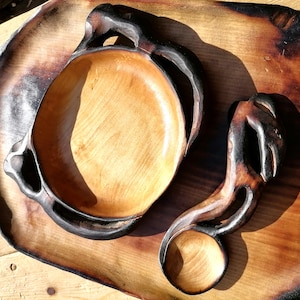May include: Hand-carved wooden bowl and spoon set. The bowl has a light wood interior and dark, textured handles. The spoon features a rounded bowl and a matching dark handle. The set rests on a wooden tray.