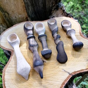 Pode incluir: Cinco colheres de madeira artesanais com designs únicos. As colheres variam em forma e cor, algumas com detalhes escuros e queimados e outras com um acabamento de madeira clara natural. Estão dispostas sobre uma fatia de madeira natural.