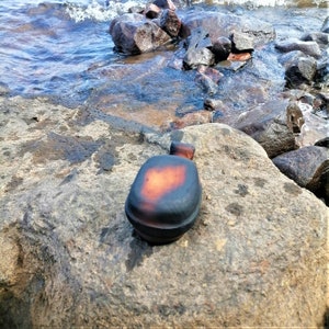 May include: A black and orange ceramic bottle with a rounded shape sits on a gray rock.