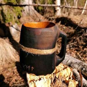 Può includere: Un boccale di legno fatto a mano con un esterno scuro tinto e un interno più chiaro. Il boccale presenta un manico ed è avvolto con spago. Simboli scolpiti e una freccia sono visibili sul davanti. Il boccale è appoggiato su una superficie di legno.