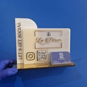 Pode incluir: Um porta-cartões de acrílico transparente com detalhes dourados. O suporte apresenta o texto "La Décor" em escrita dourada, um logotipo do Instagram e um código QR. As palavras "LET'S GET SOCIAL" são impressas verticalmente na lateral.