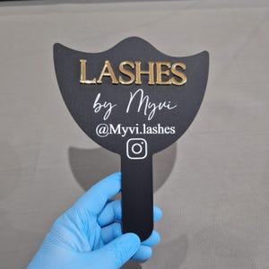 Könnte beinhalten: Ein schwarzes Handschild mit fächerförmigem Oberteil, das den goldenen erhabenen Schriftzug LASHES, den weißen Schreibschrifttext by Myvi, den weißen Text @Myvi.lashes und ein weißes Instagram-Symbol zeigt. Das Schild hat einen vertikalen Griff.