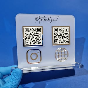 Op de afbeelding: Wit acryl bord met de tekst "Pfoten Bunt" in zwarte schrijfletters. Het bord heeft twee zwart-witte QR-codes, een zilveren Instagram-logo en een zilveren wereldbol-logo.