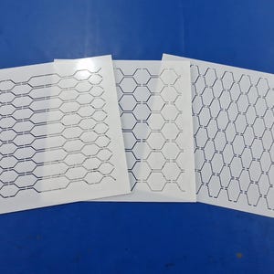 Könnte beinhalten: Drei weiße Schablonen mit geometrischen Ausschnittmustern sind auf einer blauen Oberfläche angeordnet. Die Schablonen weisen unterschiedliche Designs auf, darunter miteinander verbundene Formen und Wabenmuster. Die Schablonen werden wahrscheinlich für Bastel- oder Designprojekte verwendet.