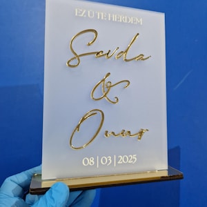Könnte beinhalten: Ein weißes Acrylschild mit goldener Aufschrift. Das Schild trägt den Text "Ez üte herdem" gefolgt von den Namen "Sevda & Onur" in Kursivschrift. Das Datum "08 | 03 | 2025" ist unter den Namen gedruckt.