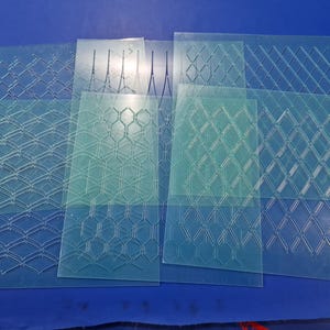 Può includere: Una collezione di stencil in plastica trasparente rettangolari con vari motivi geometrici, tra cui disegni a diamante ed esagonali. Gli stencil sono disposti su una superficie blu, mostrando la loro natura trasparente e gli intricati dettagli ritagliati.