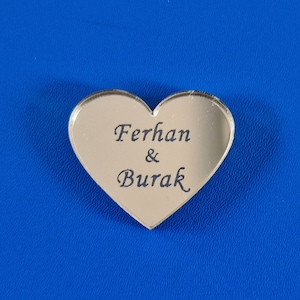 Könnte beinhalten: Ein silberner herzförmiger Spiegel mit der Inschrift "Ferhan & Burak" in schwarzen Buchstaben.