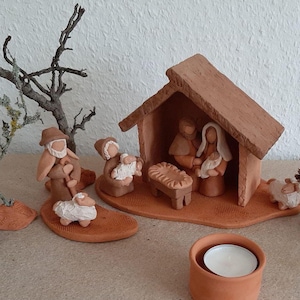 Può includere: Un presepe in terracotta con una piccola casa, figure di Maria, Giuseppe e il bambino Gesù, un pastore e una pecora. La scena è ambientata su una superficie marrone con un piccolo portacandele in terracotta.