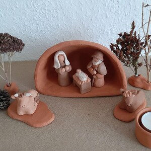 Può includere: Un presepe in terracotta con le figure di Maria, Giuseppe e il bambino Gesù. La scena include anche due animali in terracotta, un albero e un piccolo portacandele.