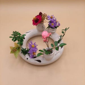 Op de afbeelding: Een witte keramische bloemenkrans met kleine geïntegreerde vazen, met levendige bloemen in tinten paars, rood en roze, en groen blad. De krans is bevestigd aan een beige muur.