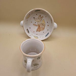 Può includere: Un set di ciotola e tazza in ceramica bianca. La ciotola presenta un'illustrazione di volpe dipinta a mano con accenti marroni e arancioni, circondata da punti e motivi a foglia. La tazza ha un manico ed è decorata con motivi a foglia simili.