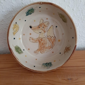Peut inclure: Un bol rond en céramique blanc cassé avec une illustration de renard peinte à la main. Le renard est brun et orange, avec une expression souriante. Le bol est décoré de feuilles vertes et marron et de petits points. Le bord est brun foncé.