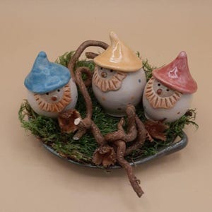 Può includere: Tre gnomi in ceramica con cappelli colorati e volti dipinti, seduti su un letto di muschio e ramoscelli in una ciotola di ceramica grigia.