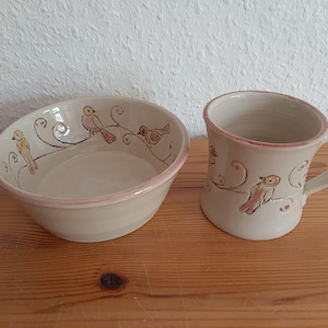 Puede incluir: Un juego de cuenco y taza de cerámica. Las piezas blanquecinas tienen un borde rosa y están decoradas con pájaros marrones sobre ramas. El cuenco es ovalado y la taza tiene un asa curvada.