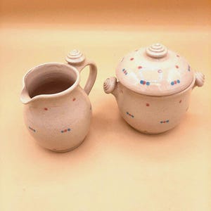 Può includere: Un set di caraffa per panna e ciotola coperta in ceramica color crema. La caraffa per panna ha un manico e un beccuccio. La ciotola ha un coperchio con un pomello. Entrambi i pezzi sono decorati con punti rossi e blu. Il set è probabilmente per servire condimenti o salse.