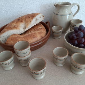 Può includere: Una natura morta con una pagnotta in un piatto di ceramica marrone, una brocca, piccole tazze e una ciotola di uva. La ceramica ha una base color crema con accenti blu e marroni. Il pane è affettato.
