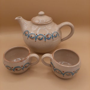 Può includere: Un set di tre tazze da tè in ceramica e una teiera, tutte di colore beige chiaro con un motivo decorativo blu e marrone.