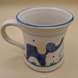 Può includere: Una tazza in ceramica bianca con un elefante blu dipinto a mano con un orecchio giallo. La tazza presenta un bordo blu, un manico curvo e piccoli punti colorati sparsi sopra l'elefante, posizionato su una sottile linea verde d'erba.