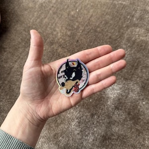 May include: A circular sticker with a black cat silhouette and a tan mouse. The sticker has a holographic finish and is held in a hand. The background of the sticker has a collage of images.