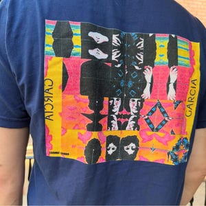May include: Navy blue t-shirt with a colorful graphic design on the back. The design features abstract shapes, faces, and the name "GARCIA" in yellow. The words "COSMIC CHAOS" are printed at the bottom.