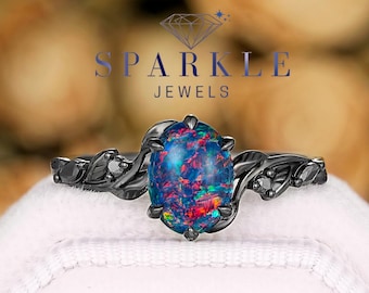 May include: A black ring with an oval-shaped, multi-colored opal gemstone. The ring features a decorative, floral design on the band and the text "SPARKLE JEWELS". The gemstone displays shades of blue, red, and green.