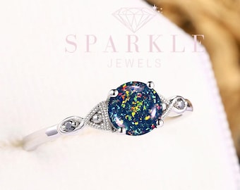 May include: A silver ring with a round, dark blue gemstone with colorful flecks. The ring has a delicate design with small accent stones on either side of the main stone. The ring is set against a white background.