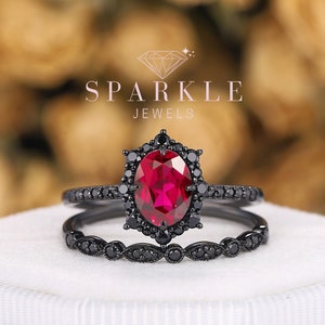 May include: Black and red engagement ring set with a large oval ruby gemstone surrounded by smaller black diamonds. The ring is set in a black metal band with a delicate design. The ring is displayed in a white box with the text "SPARKLE JEWELS" on the top.