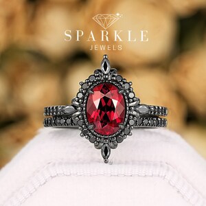 Può includere: Un set di anelli di fidanzamento neri con una grande pietra rossa ovale e pietre nere più piccole. L'anello è posto su uno sfondo sfocato di fiori gialli. Il testo "SPARKLE JEWELS" è visibile.