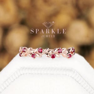 May include: A rose gold ring featuring a band designed with interconnected leaves and small round red gemstones. The text SPARKLE JEWELS is visible above the ring. The ring is positioned on a white surface with a blurred background.