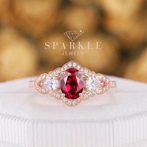 May include: Rose gold ring featuring an oval-cut ruby center stone, flanked by pear-shaped white gemstones. The ring has a detailed filigree design and is set on a white velvet surface. The ring is from Sparkle Jewels.