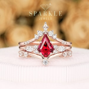 May include: A three-piece rose gold engagement ring set with a red gemstone in the center and smaller diamonds on the band. The ring is on a white box with the text "SPARKLE JEWELS" above the ring.