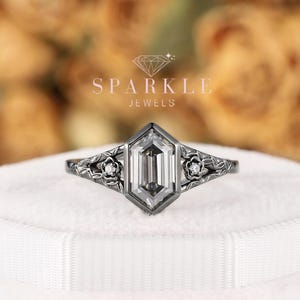 May include: A dark metal ring featuring a central hexagonal-cut clear stone. The band displays intricate floral engravings on both sides, each accented with a small, round, clear stone. The text SPARKLE JEWELS is visible above the ring.
