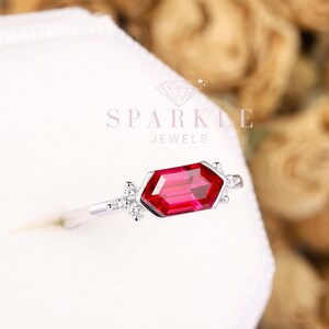 May include: A silver ring with a red octagon-shaped gemstone and small clear gemstones on the band. The ring is in a white box with the text "Sparkle Jewels" on it.
