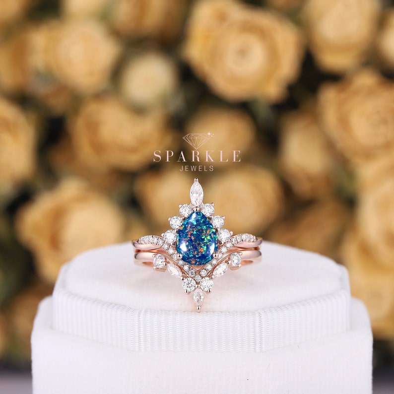 May include: A rose gold engagement ring with a pear-shaped blue opal center stone and a halo of smaller round diamonds. The ring is set on a white band and has a matching wedding band with a delicate design of diamonds.