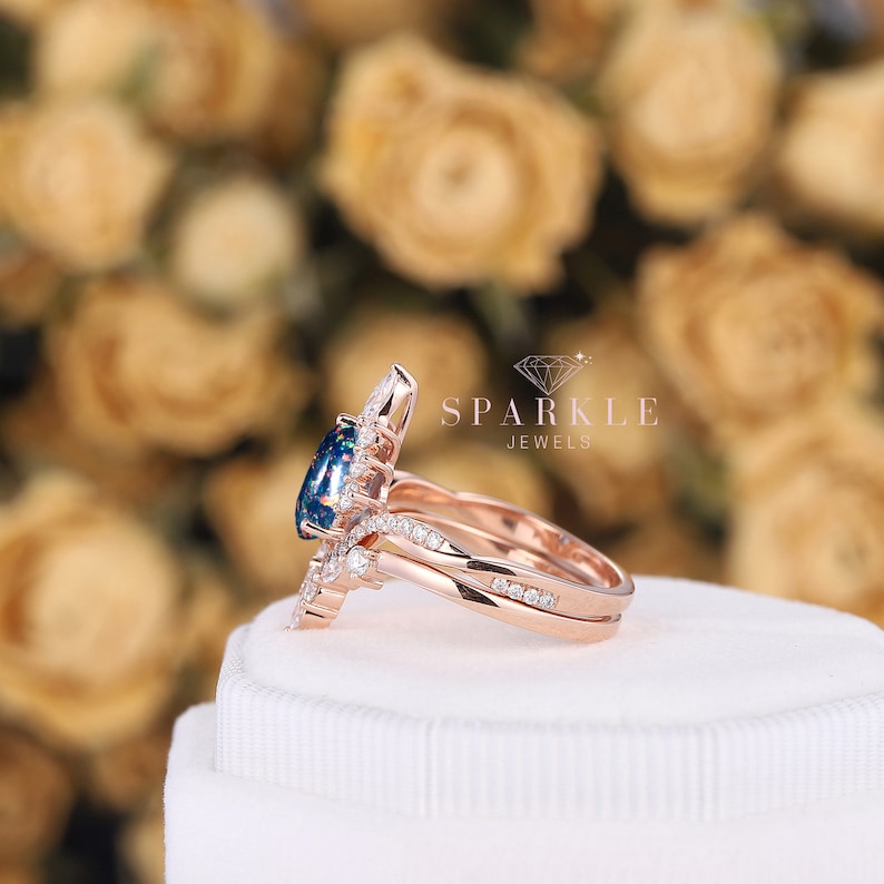 K&ouml;nnte beinhalten: Ein Verlobungsring aus Ros&eacute;gold mit einem gro&szlig;en ovalen schwarzen Opal, der von kleineren Diamanten umgeben ist. Der Ring liegt auf einer wei&szlig;en Schachtel mit dem Text "Sparkle Jewels" darauf.