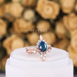 K&ouml;nnte beinhalten: Ein Verlobungsring mit einem birnenf&ouml;rmigen Opal in Ros&eacute;gold, einem zarten floralen Design und kleineren Diamanten. Der Ring liegt auf einer wei&szlig;en Schachtel mit dem Text "SPARKLE JEWELS" &uuml;ber dem Ring.
