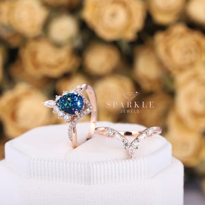 K&ouml;nnte beinhalten: Ein Paar Verlobungsringe aus Ros&eacute;gold mit einem birnenf&ouml;rmigen schwarzen Opal-Edelstein, der von kleineren Diamanten umgeben ist. Die Band ist mit einem zarten Diamantenmuster verziert. Der zweite Ring ist eine einfache Band mit einem V-f&ouml;rmigen Design, das mit kleinen Diamanten besetzt ist. Die Ringe werden in einer wei&szlig;en Schachtel mit dem Text "Sparkle Jewels" pr&auml;sentiert.
