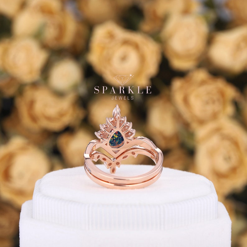 K&ouml;nnte beinhalten: Ein Ring aus Ros&eacute;gold mit einem birnenf&ouml;rmigen schwarzen Opal, der von kleineren klaren Steinen umgeben ist. Der Ring liegt auf einer wei&szlig;en Schachtel mit dem Text "SPARKLE JEWELS" &uuml;ber dem Ring.