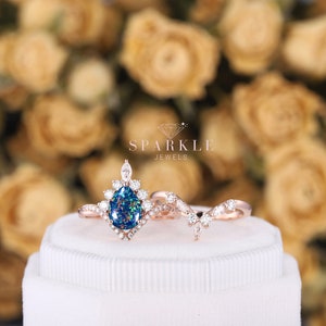 K&ouml;nnte beinhalten: Ein Verlobungsring aus Ros&eacute;gold mit einem birnenf&ouml;rmigen Opal als Mittelst&uuml;ck und einem passenden Band mit einem zarten Rankendesign. Der Ring ist in einem Halo aus kleineren Diamanten gefasst und wird auf einer wei&szlig;en Schachtel pr&auml;sentiert. SPARKLE JEWELS ist auf der Schachtel aufgedruckt.