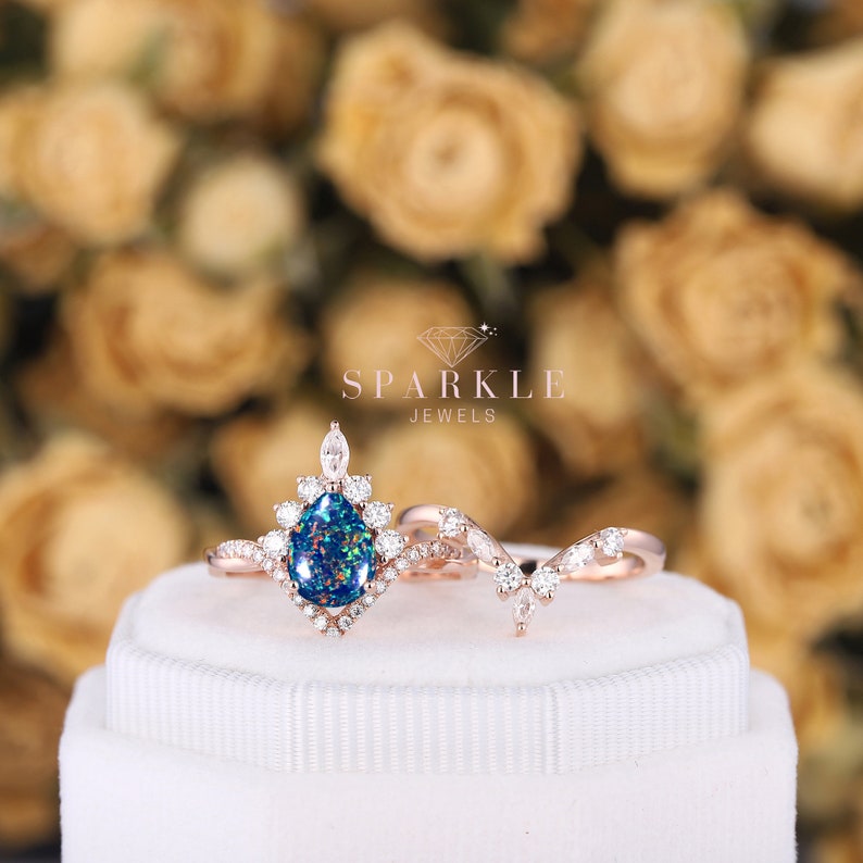 K&ouml;nnte beinhalten: Ein Verlobungsring aus Ros&eacute;gold mit einem birnenf&ouml;rmigen Opal als Mittelst&uuml;ck und einem passenden Band mit einem zarten Rankendesign. Der Ring ist in einem Halo aus kleineren Diamanten gefasst und wird auf einer wei&szlig;en Schachtel pr&auml;sentiert. SPARKLE JEWELS ist auf der Schachtel aufgedruckt.