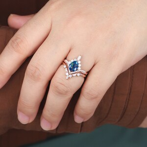K&ouml;nnte beinhalten: Ein Verlobungsring aus Ros&eacute;gold mit einem blauen, ovalen Mittelst&uuml;ck und einem Diamanten-Halo. Der Ring ist auf einem zarten Band mit kleineren Diamanten gefasst.