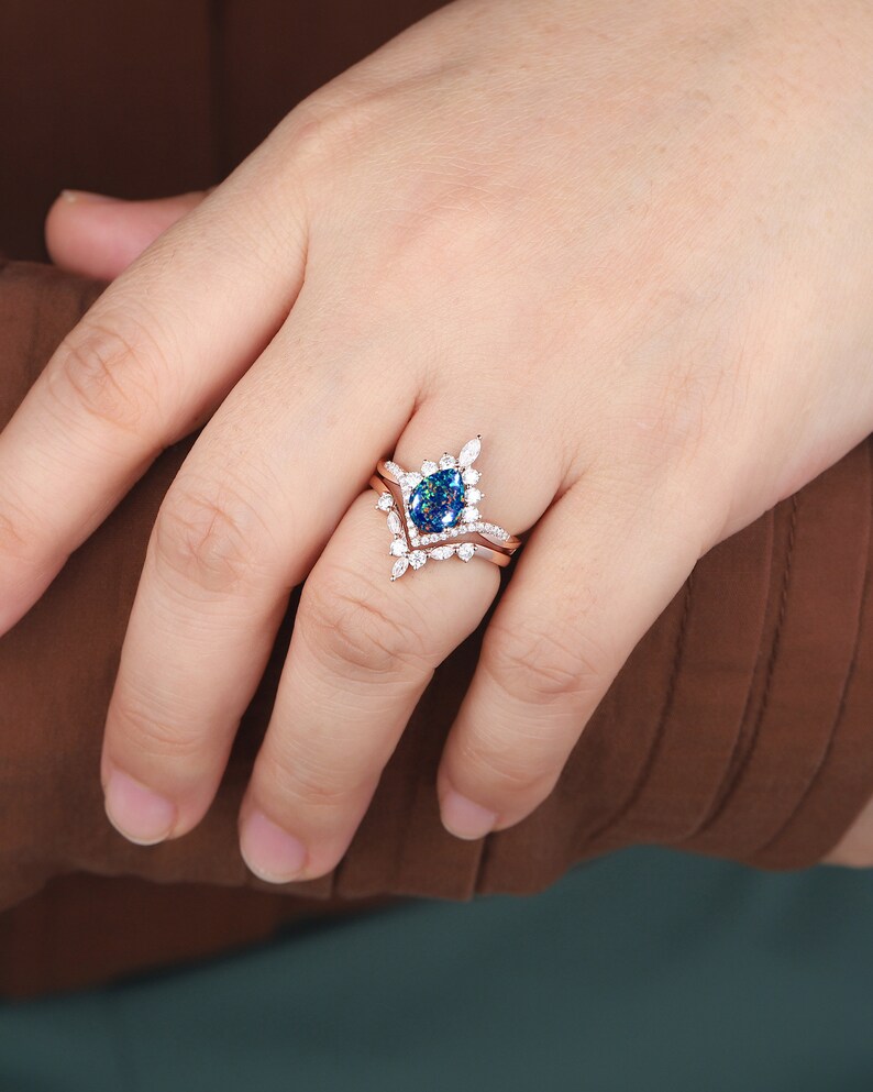 K&ouml;nnte beinhalten: Ein Verlobungsring aus Ros&eacute;gold mit einem blauen, ovalen Mittelst&uuml;ck und einem Diamanten-Halo. Der Ring ist auf einem zarten Band mit kleineren Diamanten gefasst.