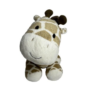 May include: A plush giraffe toy with a white face, brown eyes, and a brown nose. The giraffe is sitting with its legs tucked underneath it. The giraffe is made of soft, plush fabric and has a brown and tan patterned body.