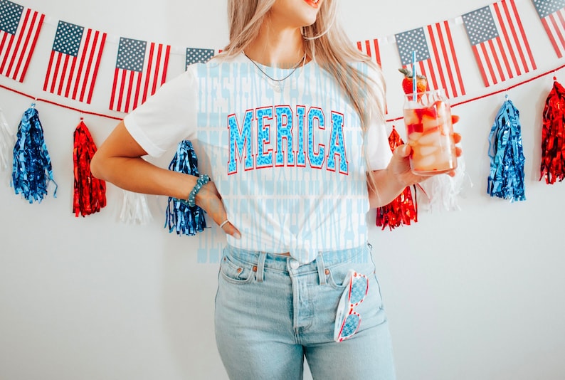 May include: A white t-shirt with a blue and red "MERICA" graphic. The shirt is worn by a person holding a glass of red liquid with a strawberry garnish. The background features a red, white, and blue string of flags and tassels.