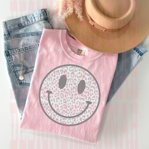 May include: Pink t-shirt with a gray smiley face design featuring a leopard print pattern. The shirt is laid out on a denim background with a brown hat.
