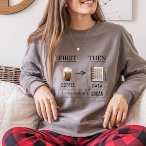 May include: A gray sweatshirt with the text "FIRST THEN COFFEE -> DATA I am earning a: BREAK". The graphic shows a coffee drink and a spreadsheet. The sweatshirt is worn with red and black plaid pajama pants.