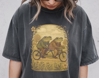 May include: Dark grey t-shirt with a charming illustration of two frogs on a tandem bicycle. The frogs wear jackets and are set against a park scene with flowers and trees. The design is framed by a floral border, creating a vintage aesthetic.