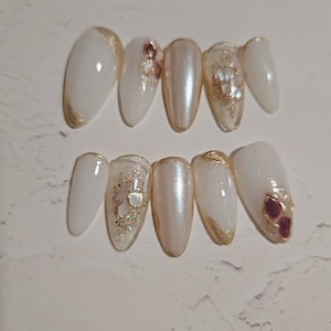 Puede incluir: Un conjunto de uñas postizas blancas en forma de almendra con detalles dorados. Algunas uñas presentan purpurina iridiscente, adornos de perlas y gemas de color burdeos. Las uñas están dispuestas sobre una superficie clara.