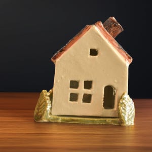 May include: A ceramic house figurine with a cream-colored body, a red-brown roof, and a brown chimney. The house has cut-out windows and a door, with green foliage on either side. The figurine is placed on a wooden surface.