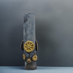 May include: A tall, cylindrical vase with a distressed, mottled gray finish. The vase is adorned with several gold-colored gear-shaped embellishments. The design has a steampunk aesthetic.